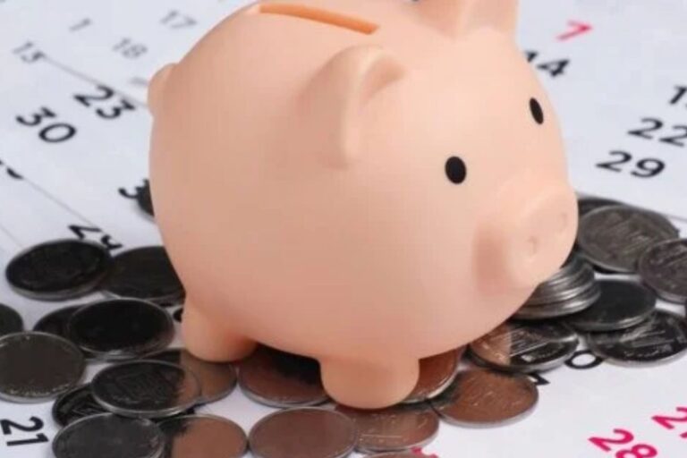 Cofrinho de porco rosa sobre uma pilha de moedas. Ao fundo, um calendário parcialmente visível com números de datas, sugerindo planejamento financeiro ou o dia de pagamento."
