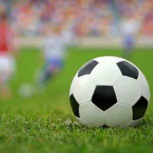 Bola de futebol em campo com jogadores ao fundo, representando a expectativa para a Copa do Mundo 2026.