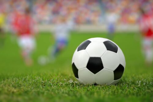 Bola de futebol em campo com jogadores ao fundo, representando a expectativa para a Copa do Mundo 2026.