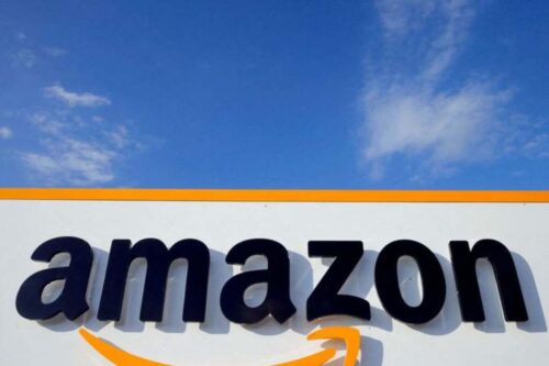 Fotografia em ângulo baixo do logotipo da Amazon em letras pretas sobre um painel branco com detalhes em laranja.