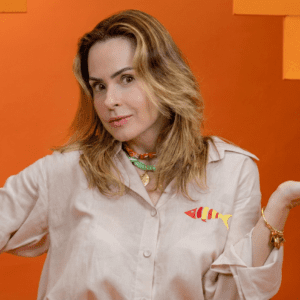 Mulher de pele clara e cabelos loiros médios ondulados, identificada como Ana Paula Renault, posando contra um fundo laranja vibrante com texturas geométricas.