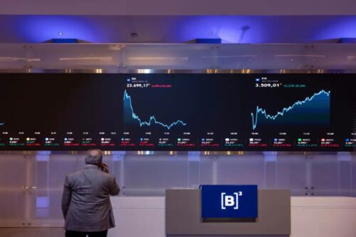 Homem de costas, ao telefone, observando um grande painel digital de cotações financeiras na sede da B3 (Bolsa de Valores do Brasil).