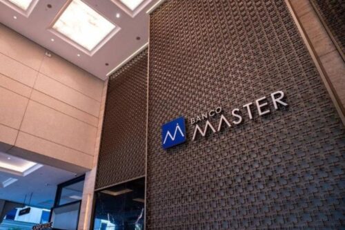 Fachada moderna do Banco Master em um ambiente corporativo. O logotipo, composto por um 'M' estilizado em um quadrado azul ao lado do nome 'BANCO MASTER' em letras prateadas, está fixado em uma parede metálica decorativa com padrão vazado.