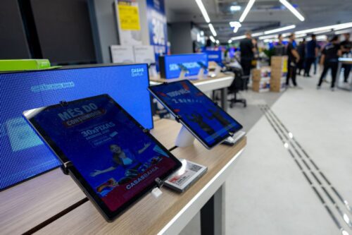 Primeiro plano de tablets exibindo anúncios da Casas Bahia em uma loja de varejo iluminada.