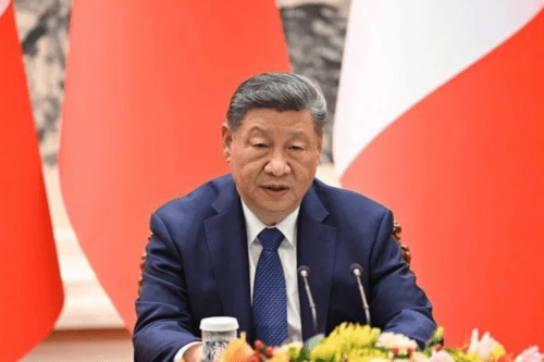 O presidente da China, Xi Jinping, sentado em uma mesa oficial com microfones à sua frente. Ele veste um terno azul-marinho, camisa branca e gravata azul texturizada. Ao fundo, aparecem bandeiras nas cores vermelho e branco. Xi Jinping está com uma expressão serena e profissional.