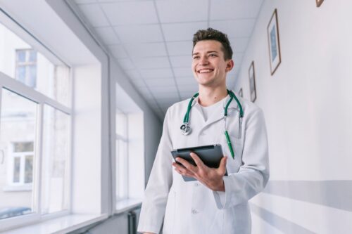 Médico jovem e sorridente caminhando pelo corredor de um hospital, vestindo jaleco branco e estetoscópio verde no pescoço, segurando um tablet digital.
