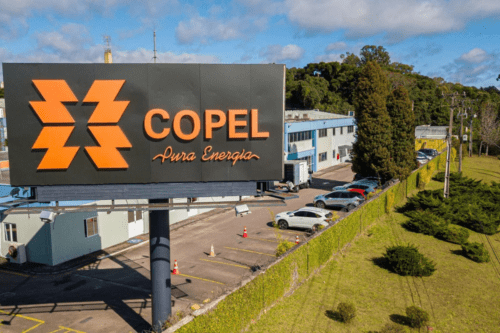 Imagem de uma usina de energia da Copel com logo destacado e estacionamento ao fundo, cercado por áreas verdes e árvores, sob céu com nuvens.