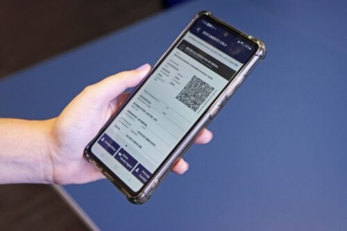 Uma mão segura um celular inclinado exibindo o documento veicular digital (CRLV) no Rio de Janeiro. A interface do aplicativo mostra detalhes do veículo e botões de ação na parte inferior. O fundo é uma superfície azul sólida e uniforme.
