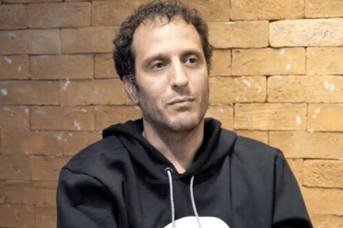 Retrato de João Ricardo Mendes, ex-CEO do Hurb (Hotel Urbano). Ele é um homem de pele clara, cabelos escuros e levemente cacheados, vestindo um casaco de moletom preto com capuz. Ele olha para o lado com uma expressão séria. Ao fundo, uma parede de tijolos aparentes em tons de bege e marrom.