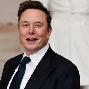 Fotografia em plano médio de Elon Musk sorrindo, vestindo um paletó preto e gravata azul escura.