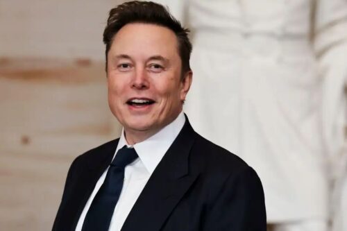 Fotografia em plano médio de Elon Musk sorrindo, vestindo um paletó preto e gravata azul escura.