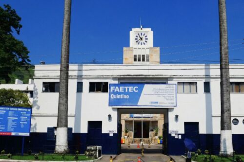 Fachada frontal do prédio branco da FAETEC Quintino sob um céu azul limpo.