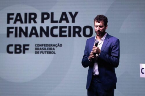 Homem de terno azul segura um microfone e fala durante um evento. Ao fundo, um painel exibe em letras grandes "FAIR PLAY FINANCEIRO" e o logotipo da CBF (Confederação Brasileira de Futebol).