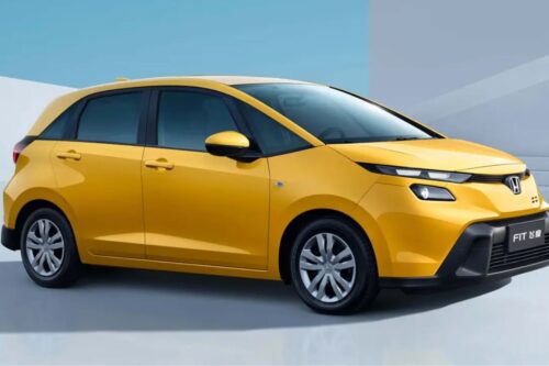 Fotografia de um carro Honda Fit de cor amarela vibrante, posicionado de perfil em um estúdio com fundo azul e branco. O veículo apresenta design moderno, faróis em LED afilados e rodas de liga leve prateadas.