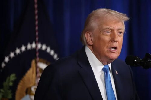 Fotografia de Donald Trump falando ao microfone durante um pronunciamento oficial, com fundo azul marinho e a bandeira dos Estados Unidos parcialmente visível ao fundo.