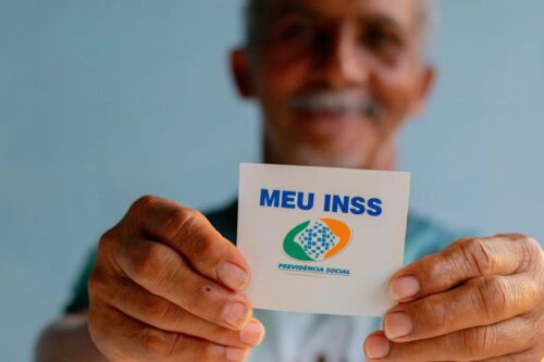 Close-up das mãos de um homem idoso segurando um cartão branco com a inscrição 'MEU INSS' e a logomarca da Previdência Social.