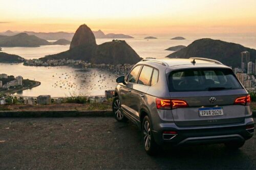 Vista traseira de um SUV prata estacionado em um mirante com o Pão de Açúcar e a Baía de Guanabara ao fundo durante o pôr do sol. A placa do veículo exibe o texto "IPVA 2026", simbolizando o imposto automotivo no contexto brasileiro.