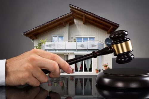 Mão de um leiloeiro segurando um martelo de madeira (gavel) prestes a bater na base, com a imagem de uma casa moderna de dois andares ao fundo. A composição simboliza um leilão de imóveis.
