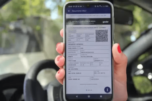 Mão de uma pessoa segurando um smartphone dentro de um carro. A tela do celular exibe o CRLV-e (Certificado de Registro e Licenciamento de Veículo digital), com o logotipo do Governo Federal e um QR Code visível. Ao fundo, é possível ver o volante e o painel do veículo de forma desfocada, reforçando o contexto de uso do documento digital no dia a dia.
