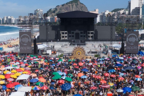 Multidão com guarda-sóis coloridos na areia da Praia de Copacabana em frente a um grande palco de evento musical, com montanhas e a orla do Rio de Janeiro ao fundo.