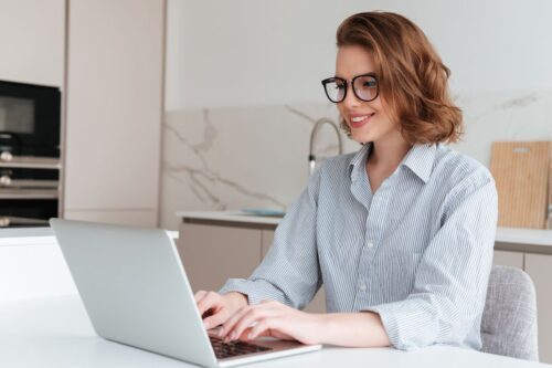 Mulher jovem com óculos e camisa listrada sorri enquanto trabalha em um notebook prata.