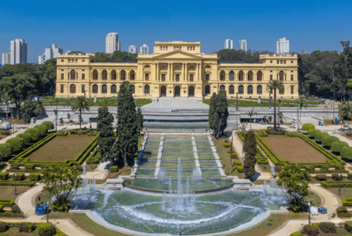 Fotografia aérea da fachada principal do Museu do Ipiranga, em São Paulo, um imponente edifício neoclássico de cor amarela clara.