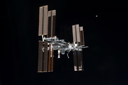 Uma imagem da Estação Espacial Internacional (ISS) flutuando na vastidão negra do espaço, com a Lua visível ao fundo como um pequeno ponto brilhante.