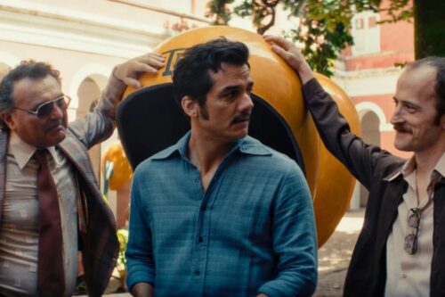 Três homens de meia-idade, com vestimentas e estilos que remetem aos anos 70 ou 80, estão posicionados ao redor de um orelhão (telefone público) amarelo clássico em uma praça ao ar livre.