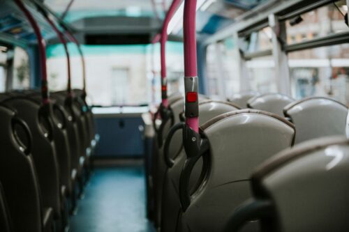 Visão do corredor interno de um ônibus urbano vazio, com fileiras de assentos cinzas e corrimãos verticais na cor rosa vibrante, equipados com botões de parada vermelhos.