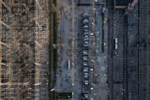 Vista aérea zenital de uma subestação de energia elétrica. A imagem mostra a complexa rede de torres de transmissão, transformadores e cabos metálicos organizados em linhas paralelas sobre um terreno industrial acinzentado.