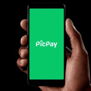 Uma mão segura um smartphone posicionado verticalmente contra um fundo preto. A tela do celular exibe um fundo verde sólido com o logotipo branco da "PicPay" centralizado.