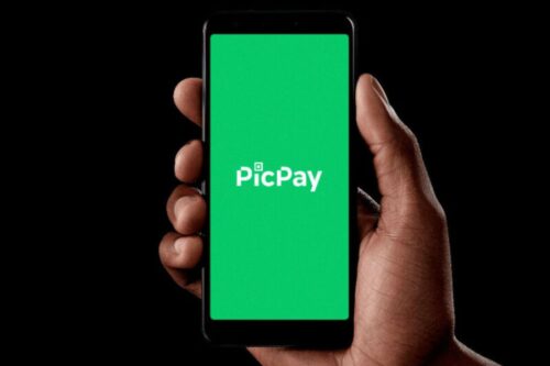 Uma mão segura um smartphone posicionado verticalmente contra um fundo preto. A tela do celular exibe um fundo verde sólido com o logotipo branco da "PicPay" centralizado.