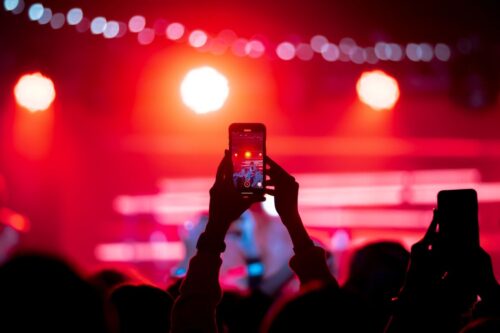 Pessoa segurando um smartphone para gravar um show ao vivo em um palco iluminado por luzes vermelhas intensas. A plateia aparece em silhueta ao fundo.