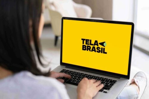 Uma mulher vista de costas utiliza um notebook que exibe em destaque o logotipo "TELA BRASIL" em letras pretas sobre um fundo amarelo vibrante.