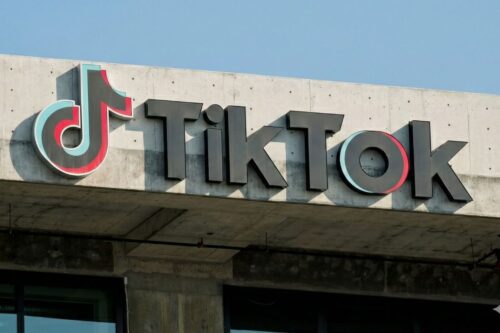 Logotipo do TikTok em letras pretas tridimensionais fixado em uma estrutura de concreto cinza sob um céu azul claro.