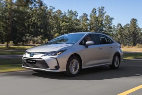 Fotografia de um Toyota Corolla Cross XRX Hybrid 2025 prata em movimento em uma estrada asfaltada, com árvores ao fundo sob luz solar.