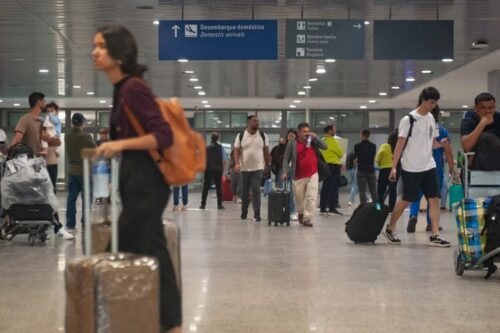 Passageiros e turistas circulam pelo saguão de um aeroporto carregando malas de rodinha e mochilas.