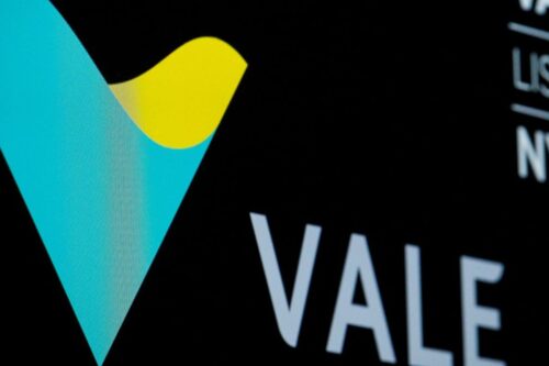 Detalhe em close do logotipo da mineradora Vale. A imagem mostra o ícone em formato de 'V' estilizado nas cores verde e amarelo sobre um fundo preto fosco. Abaixo do ícone, parte da palavra 'VALE' aparece escrita em letras brancas e modernas.