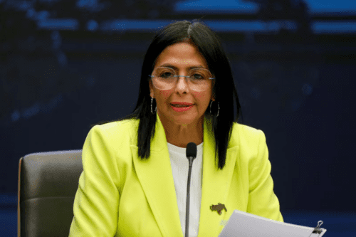 Fotografia de Delcy Rodríguez, vice-presidente da Venezuela. Ela tem cabelos pretos e lisos, usa óculos de grau e veste um blazer verde-limão vibrante sobre uma blusa branca. Delcy está sentada atrás de um microfone em um ambiente oficial, segurando papéis e olhando para a frente com uma expressão séria.