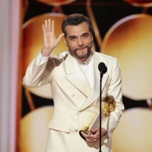 O ator brasileiro Wagner Moura em um palco de premiação, vestindo um smoking branco e segurando um troféu dourado enquanto acena para o público com a mão direita.