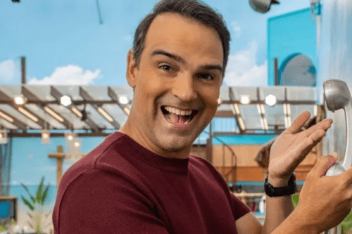 Retrato em close do apresentador Tadeu Schmidt sorrindo e olhando para a câmera. Ele veste uma camiseta bordô e segura o telefone prateado (o 'Big Fone') do reality show Big Brother Brasil. O fundo mostra a área externa da casa com céu azul e decoração do programa.