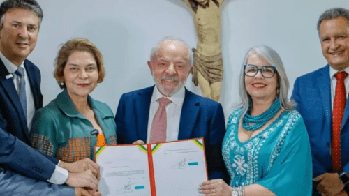 Lula aparece ao centro segurando documento assinado, ao lado de autoridades, durante cerimônia oficial em ambiente institucional.