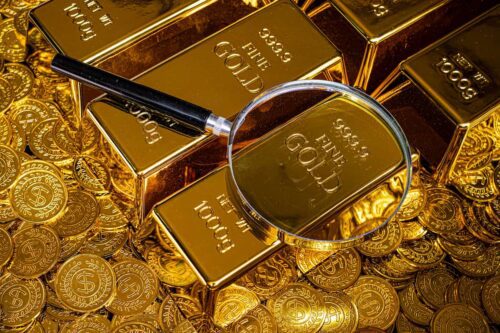 Barras de ouro empilhadas sobre moedas douradas, com lupa destacando a inscrição “gold” em uma das barras.