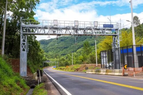Pórtico de pedágio free flow na Dutra sobre rodovia em área verde, com pista asfaltada, sinalização e estrutura metálica.