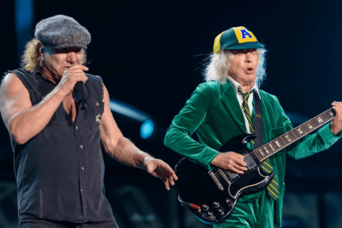Brian Johnson canta em um microfone vestindo colete preto e boina cinza, ao lado de Angus Young, que toca sua guitarra Gibson SG usando seu icônico uniforme escolar verde e boné.