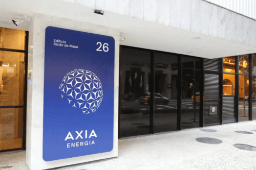 Fachada de um edifício comercial onde se destaca uma placa vertical azul da empresa Axia Energia. A placa contém um logotipo branco em formato de esfera estilizada composta por padrões geométricos, o nome "AXIA ENERGIA" na parte inferior e a identificação "Edifício Barão de Mauá, 26" no topo. O prédio possui arquitetura moderna com acabamento em mármore claro, grandes janelas de vidro escuro e calçada de pedras portuguesas. Ao fundo, é possível ver o reflexo de carros e da rua no vidro.
