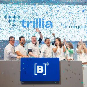 Fotografia comemora o lançamento da Trillia na B3. Um grupo de executivos sorridentes, vestindo camisas polo brancas, está posicionado atrás de um balcão cinza com o logotipo iluminado da B3.