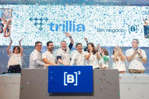 Fotografia comemora o lançamento da Trillia na B3. Um grupo de executivos sorridentes, vestindo camisas polo brancas, está posicionado atrás de um balcão cinza com o logotipo iluminado da B3.
