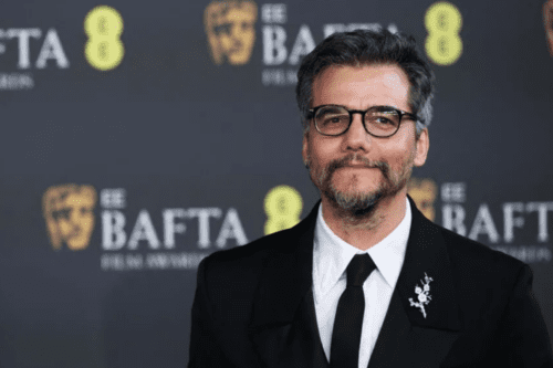 O ator brasileiro Wagner Moura posa no tapete vermelho do BAFTA Film Awards. Ele está de frente para a câmera, sorrindo levemente, usa óculos de armação escura, barba grisalha aparada e veste um terno preto clássico com camisa branca, gravata preta e um broche de flor prateada na lapela. O fundo é cinza escuro com os logotipos dourados e brancos da premiação "EE BAFTA"