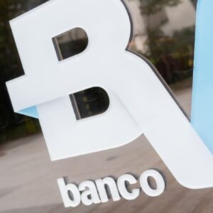 Logotipo em relevo do Banco BV fixado em uma placa de vidro transparente.